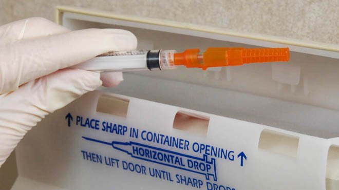 Syringe disposal. Gloved hand disposing of syringe in sharps container. Difficult waste.